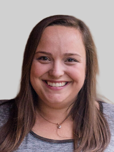 Smiling woman with long brown hair wearing a gray top and pendant necklace